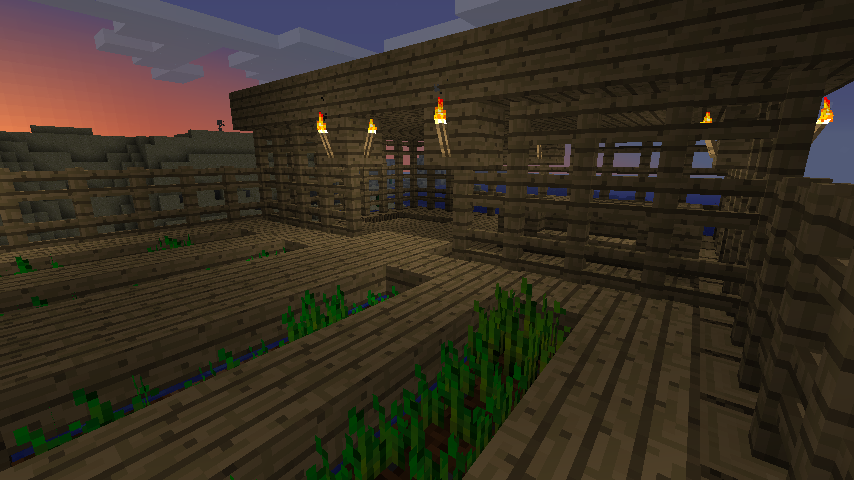 A large, unfinished building with a wooden floor and walls, featuring a row of torches along the top. The building is situated in a grassy area with a few trees nearby.